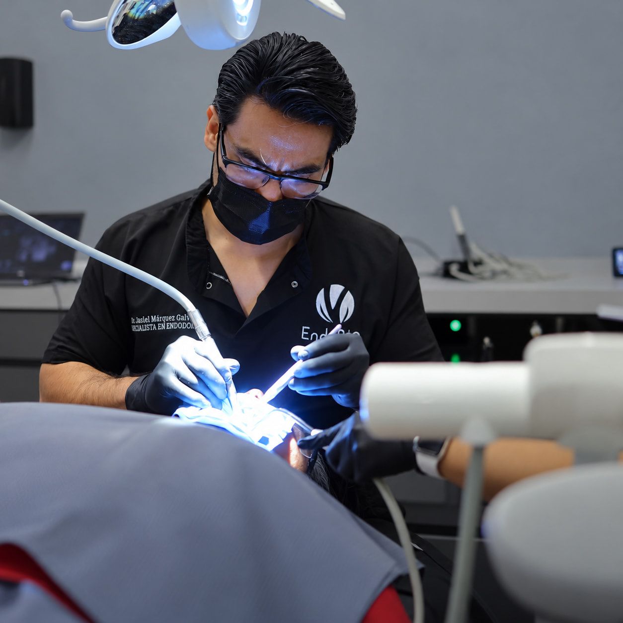 El doctor, con lentes y cubrebocas negro, atiende a un paciente bajo la lámpara del sillón dental