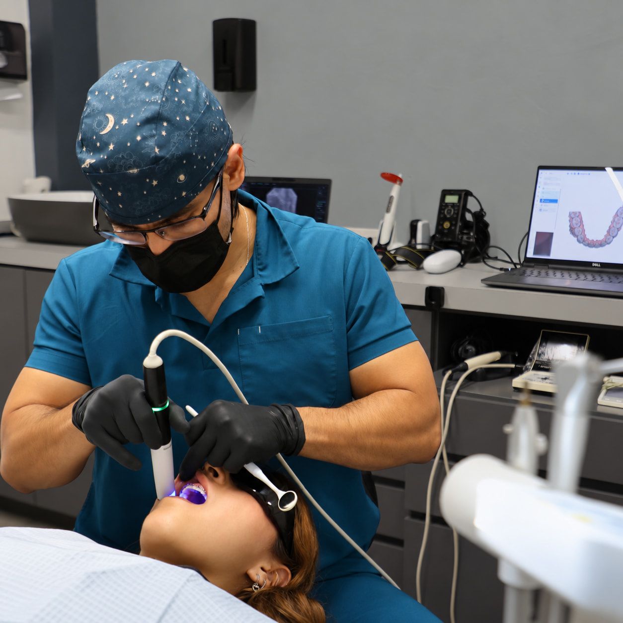 El Dr. Jasiel Márquez Galván, con gorro quirúrgico y uniforme azul, revisa a una paciente con un escáner intraoral; al fondo, una laptop muestra el modelo digital de sus dientes